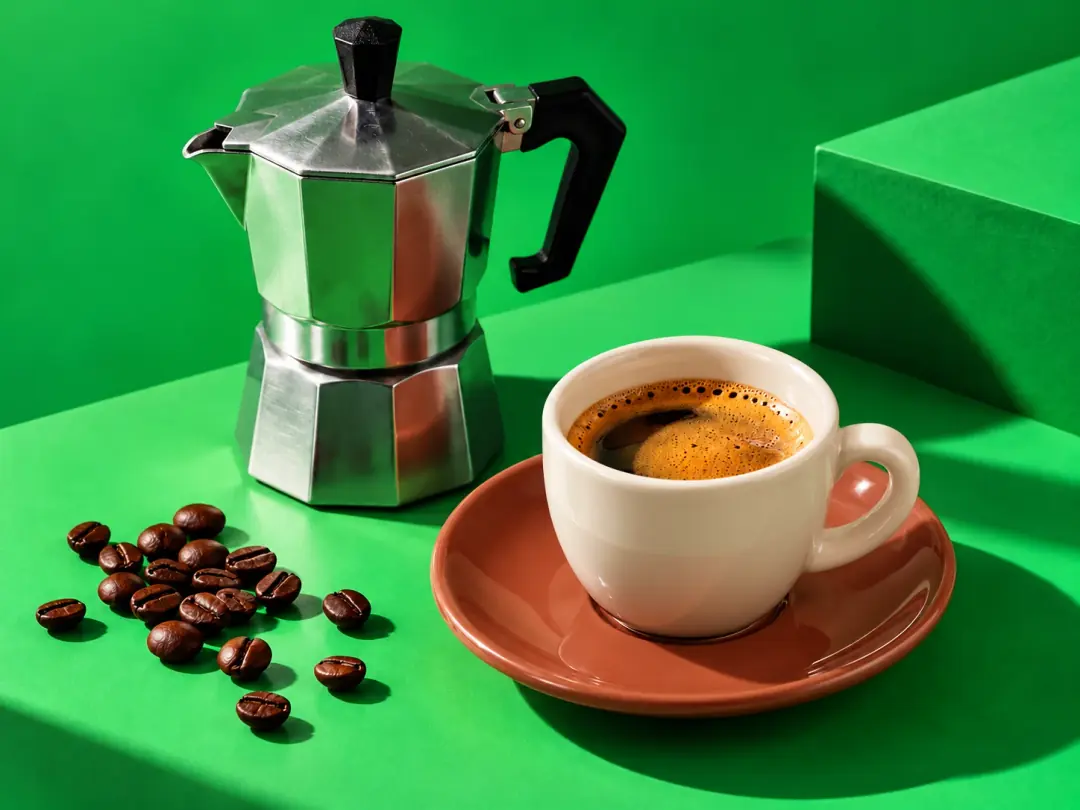 Yeşil zemin üzerinde kırık beyaz kahve fincanı, kiremit tonunda tabak ve moka pot